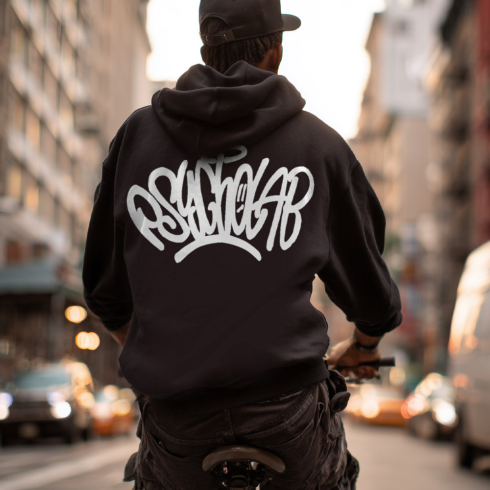 Mann auf Fahrrad trägt den Hoodie Tag Black von PsychoLab mit auffälligem Logo auf der Rückseite, umgeben von urbanem Ambiente.
